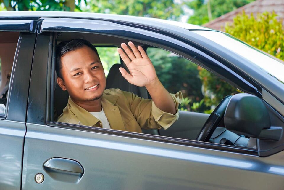 A man sitting in the driver’s seat of a car smiles and waves through the open window, with lush greenery in the background—a welcoming Bali private driver ready for your adventure.
