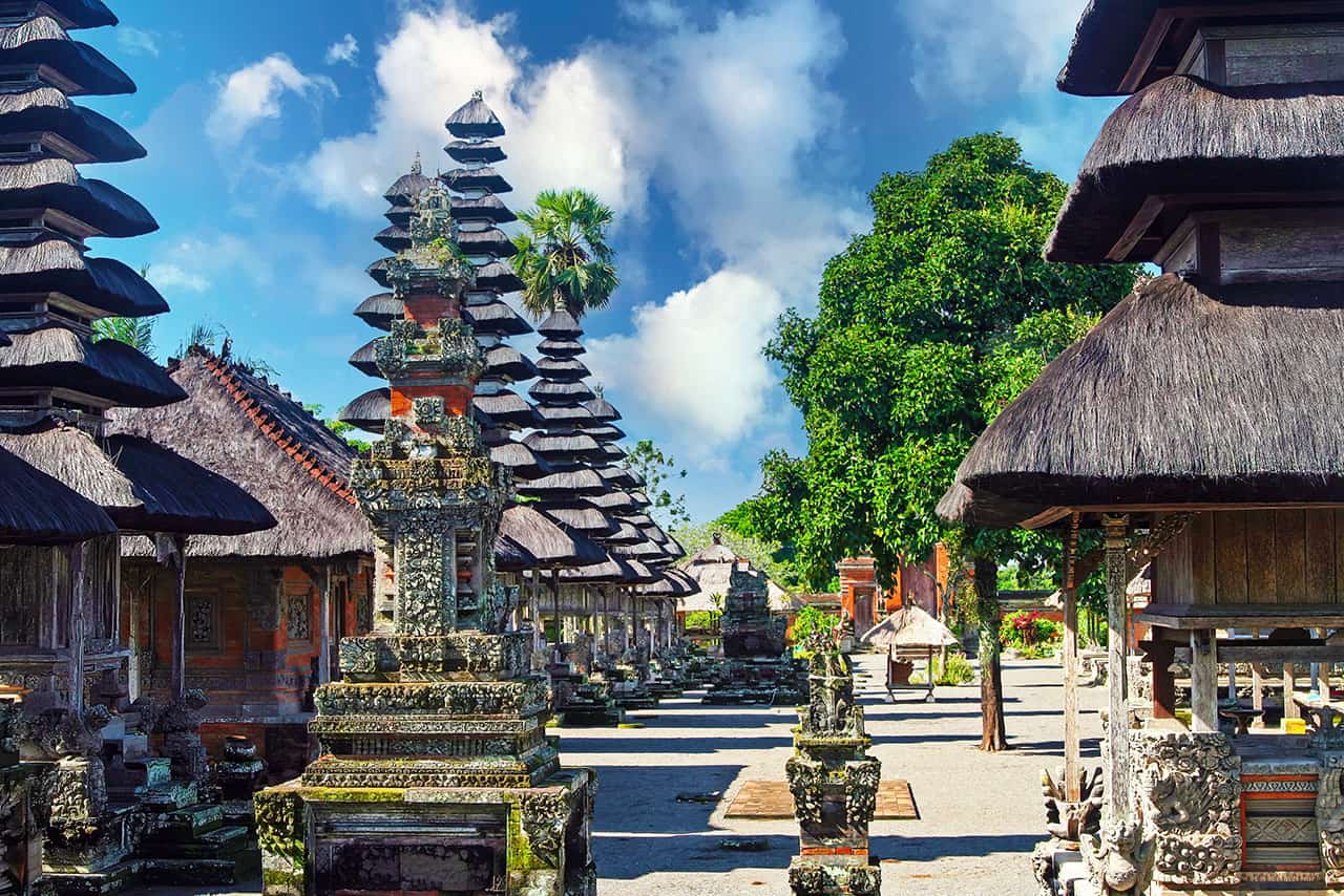 A row of traditional Balinese temples at Bali Pura Taman Ayun, featuring multi-tiered thatched roofs, ornate stone carvings, and lush green trees under a bright blue sky with scattered clouds.