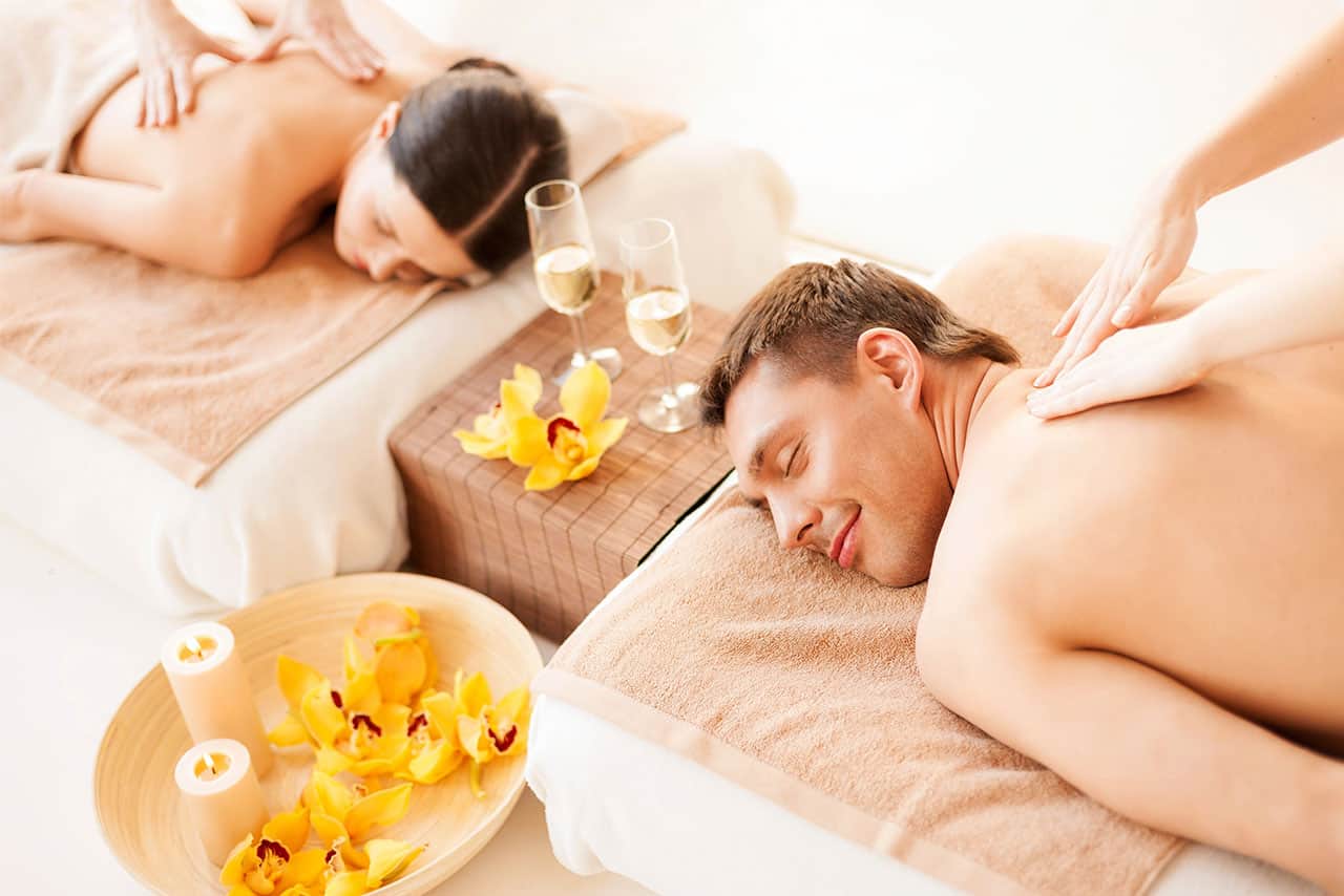A man and a woman lie side by side on massage tables, receiving back massages. Surrounded by yellow flowers, candles, and two glasses of wine, they enjoy a relaxing Bali honeymoon spa atmosphere.