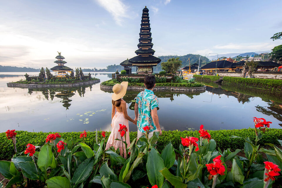 A couple stands by a pond with red flowers, gazing at the Danu Beratan Bali pagoda temple, surrounded by water and lush greenery beneath a blue sky.