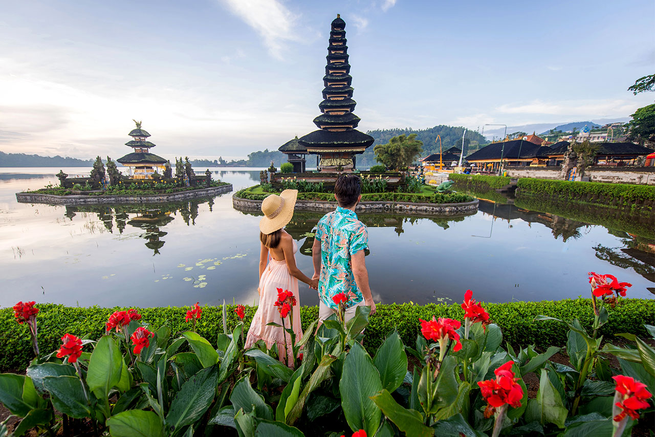 A couple stands by a pond with red flowers, gazing at the Danu Beratan Bali pagoda temple, surrounded by water and lush greenery beneath a blue sky.