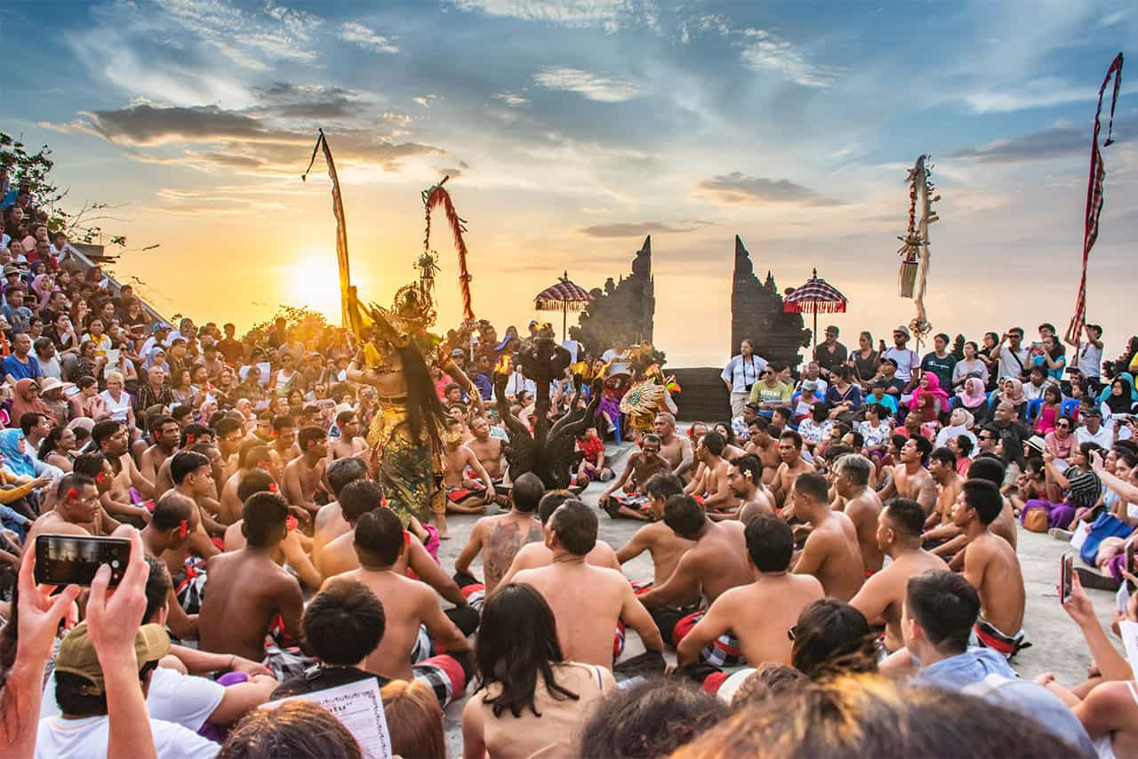 A large crowd watches a captivating kecak dance performance at sunset, with dancers in sarongs seated in a circle, framed by ornate temple gates and the sea in the background.