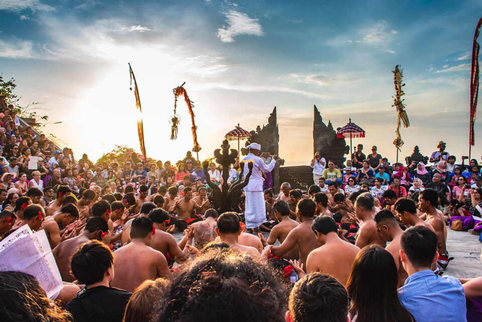 A large group gathers outdoors at sunset for a traditional Balinese ceremony. Men sit in a circle, some topless, as part of the captivating Bali Kecak Dance, while a priest in white robes stands in the center amid ornate decorations and temple gates.
