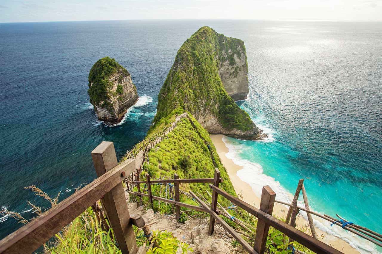 Wooden stairs descend toward a sandy beach and turquoise waters at Nusa Penida, with steep, lush green cliffs and rock formations rising dramatically from the ocean at Kelingking Beach, Indonesia.