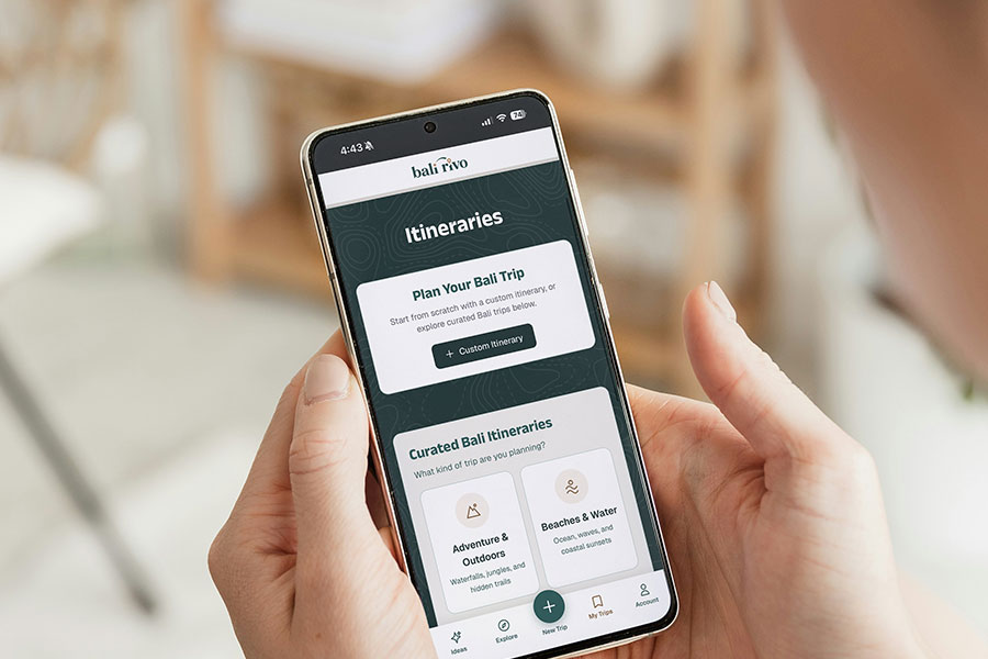 A person holds a smartphone displaying the Bali Rivo Itineraries page, featuring options to plan a Bali trip and view curated itineraries like “Adventures & Outdoors” and “Beaches & Water.”.