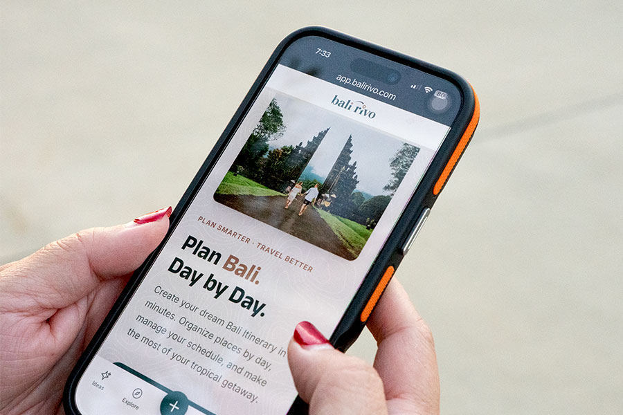 A hand holding a smartphone displaying a Bali Itinerary Builder website, featuring a photo of a temple entrance and the text "Plan Bali. Day by Day." with detailed itinerary options below.