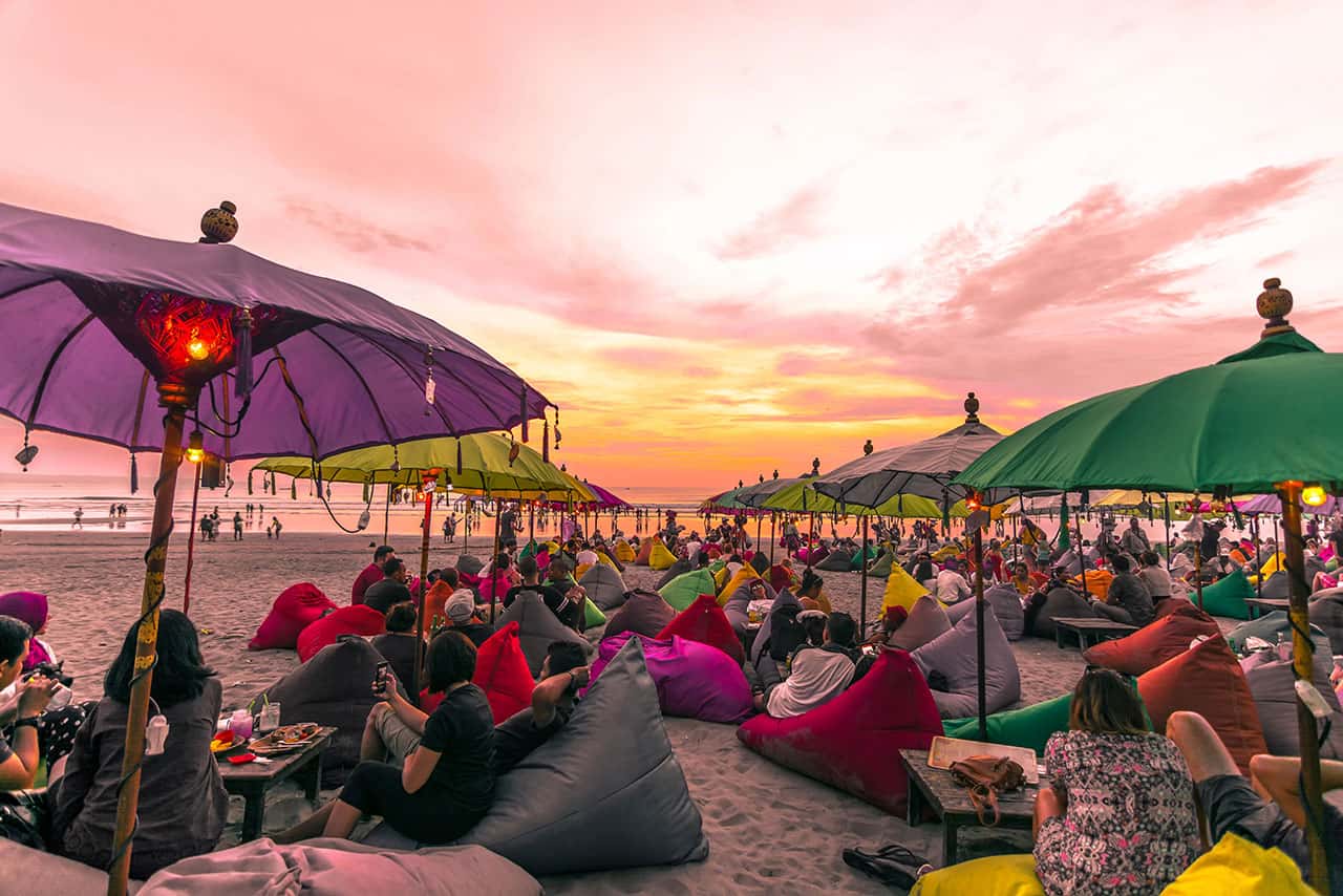 People relax on colorful beanbags under large umbrellas on the sandy shores of Seminyak Double Six Beach at sunset, enjoying the vibrant sky and lively social atmosphere as groups dine and watch the day fade away.