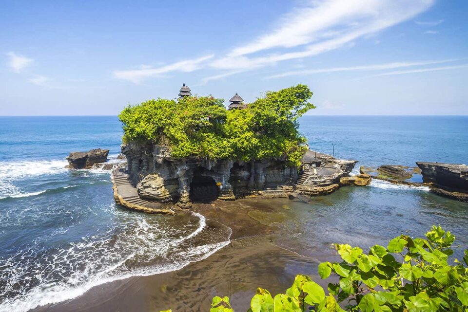 Tanah Lot temple sits atop a rocky outcrop surrounded by the ocean in Bali, Indonesia, with lush green foliage and blue sky above. Waves crash gently against the rocks at Tanah Lot's iconic seaside setting.