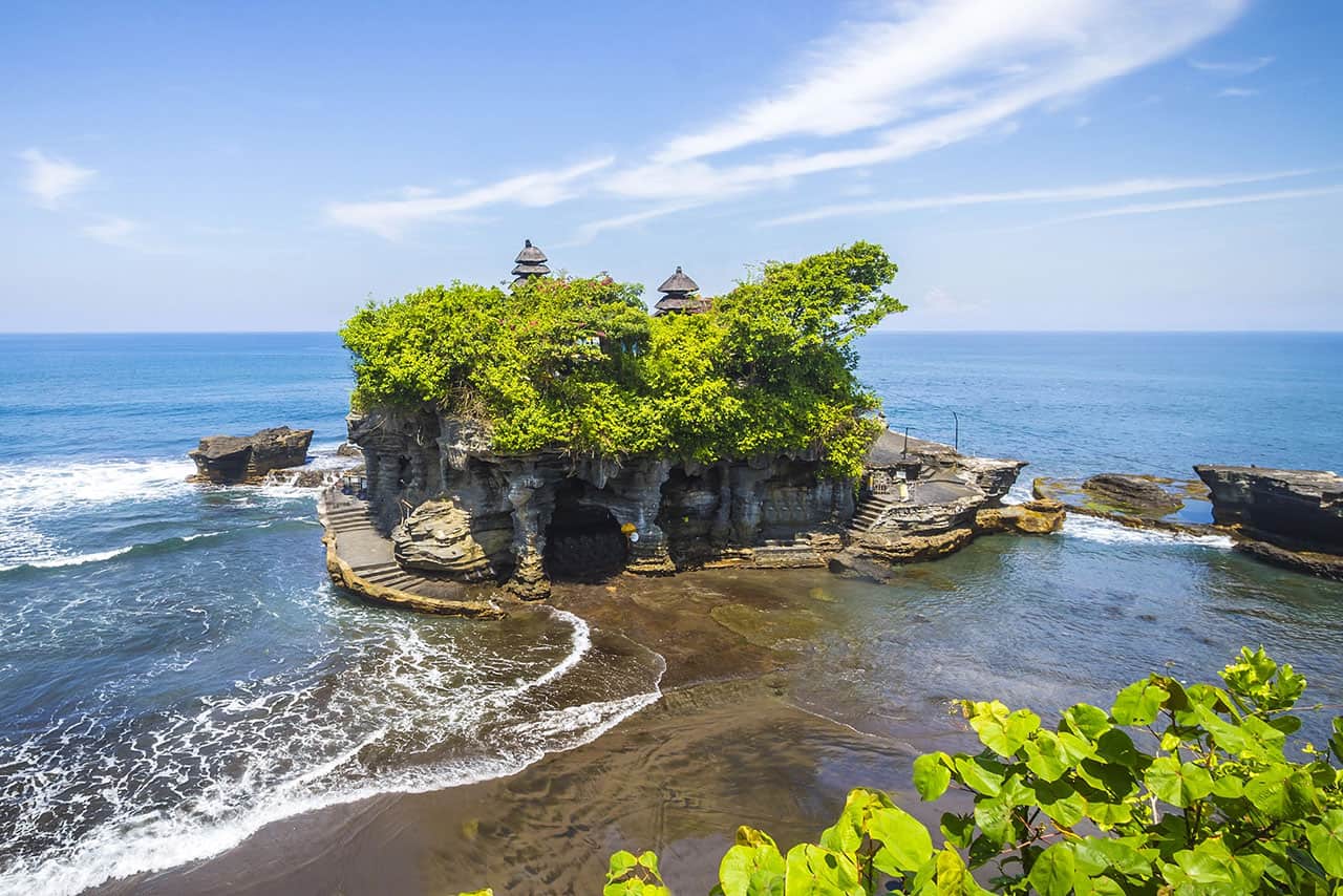 Tanah Lot temple sits atop a rocky outcrop surrounded by the ocean in Bali, Indonesia, with lush green foliage and blue sky above. Waves crash gently against the rocks at Tanah Lot's iconic seaside setting.