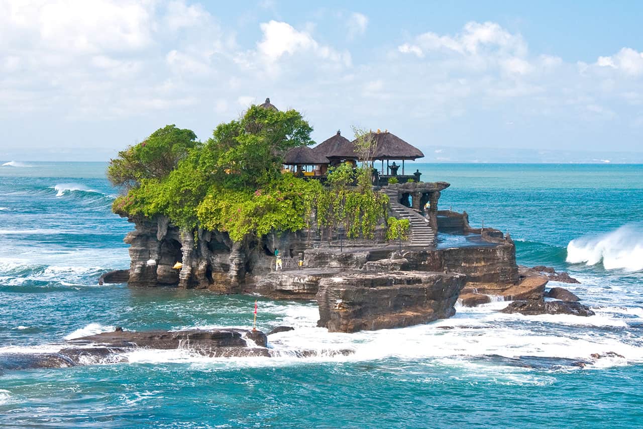 A small rocky island with lush greenery and a traditional thatched-roof temple, like Tanah Lot, sits in the turquoise sea, surrounded by waves under a partly cloudy sky.