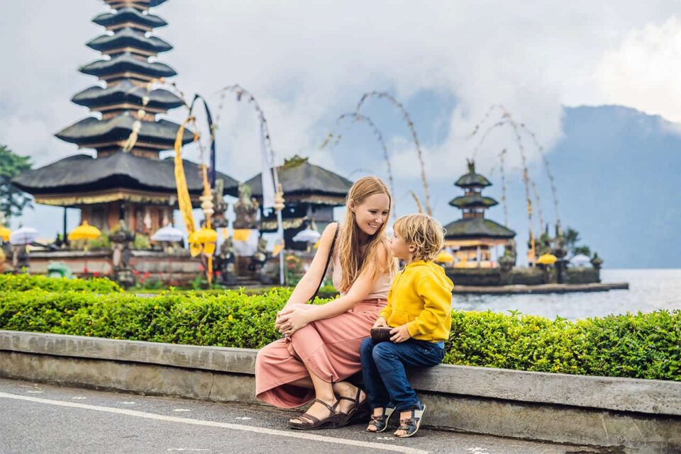A woman and a young child sit together near a road, smiling at each other, enjoying Bali with family. Traditional Balinese temples and decorated arches by a lake grace the background, with misty mountains visible in the distance.