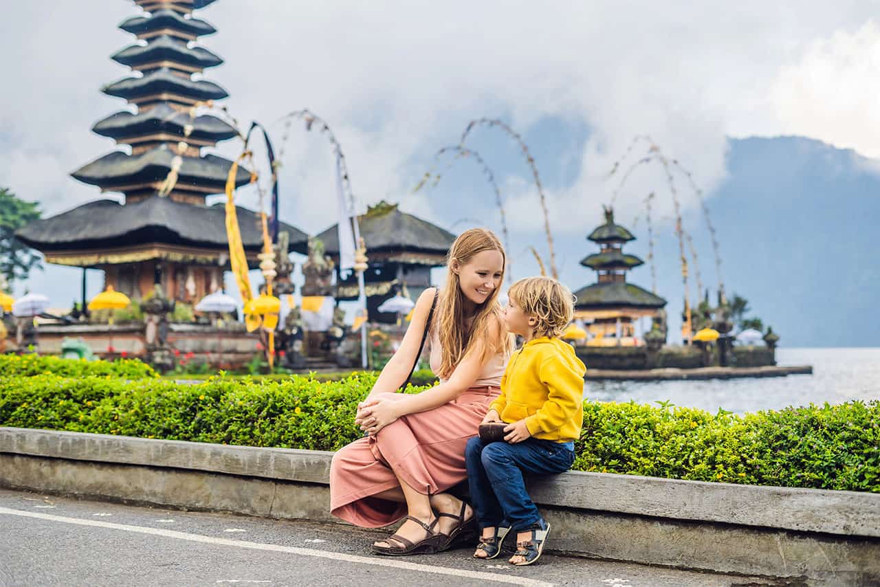 A woman and a young child sit together near a road, smiling at each other, enjoying Bali with family. Traditional Balinese temples and decorated arches by a lake grace the background, with misty mountains visible in the distance.